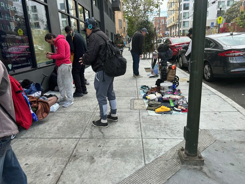 Several people are gathered on a city sidewalk next to a building; various personal items and clothing are laid out on the ground.
