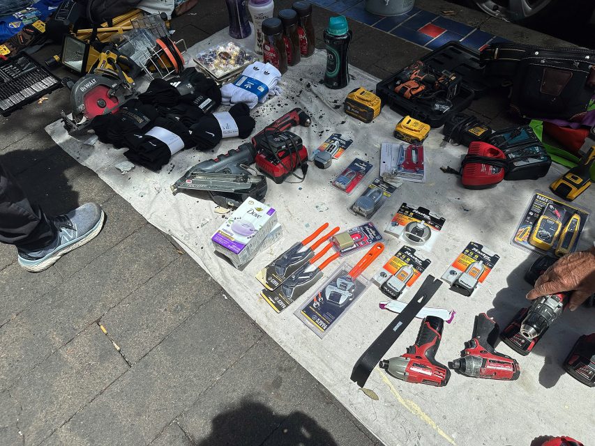 A variety of tools, batteries, flashlights, tape, and packaged items are displayed for sale on a white cloth at an outdoor market.