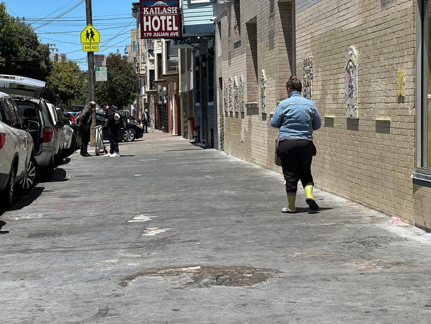 A person walks down a city sidewalk near parked cars, while two others stand by a pole under a hotel sign on a sunny day.