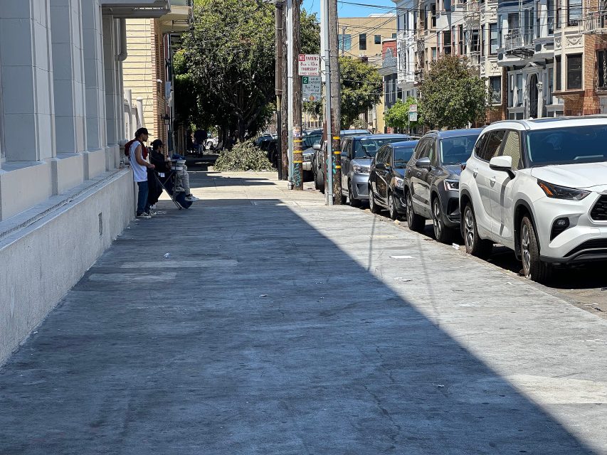 Two people stand and sit against a building on a city sidewalk; parked cars line the street, and trees cast shadows on the pavement.