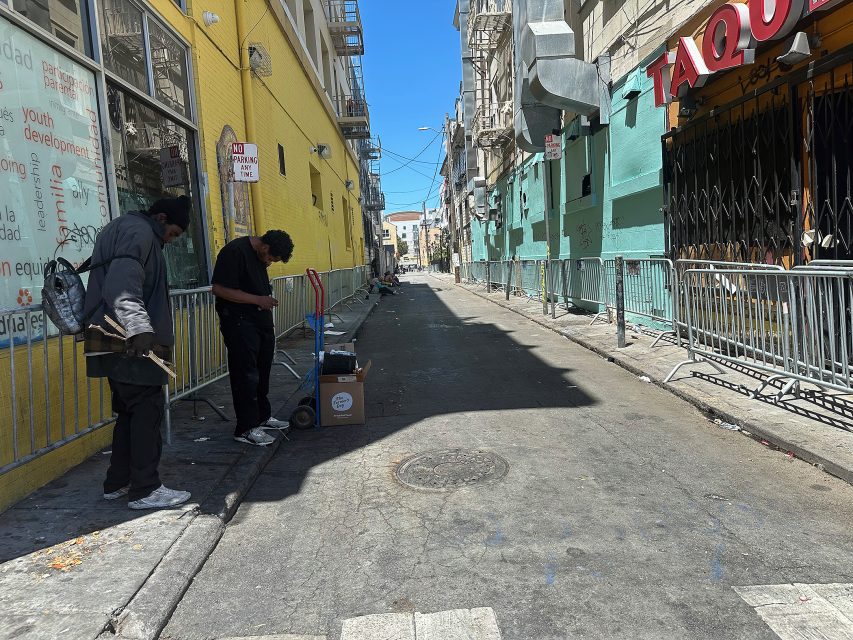 Two people stand near a box on a cart in a narrow city alleyway lined with metal barricades and colorful buildings under a clear sky.