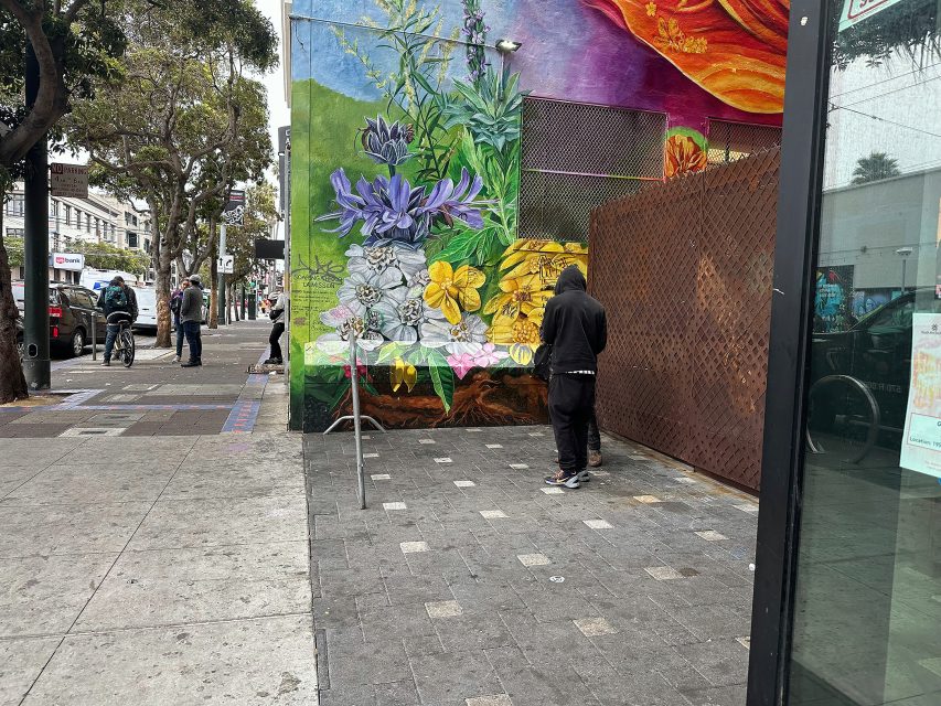 A person in a black hoodie stands near a colorful floral mural on a city sidewalk; other people and parked cars are visible in the background.