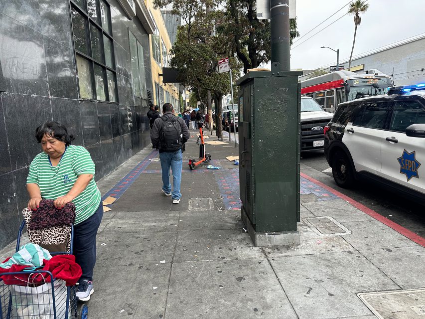 A woman pushes a cart on a city sidewalk as people walk by, with a police car parked nearby and a public transit bus in the background.