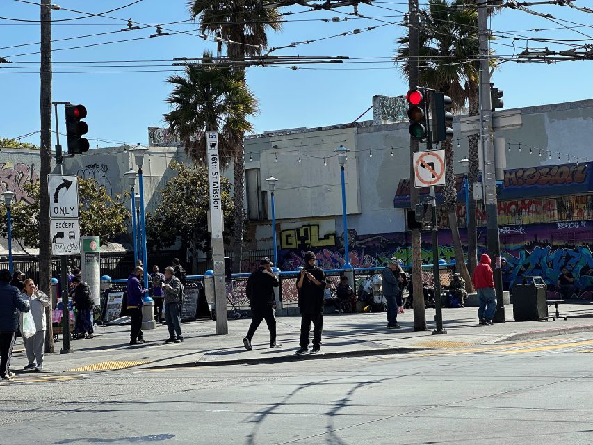 Busy urban street corner with pedestrians, graffiti-covered walls, palm trees, traffic lights, and overhead wires under a clear sky.