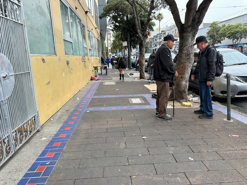 Two men stand talking on a city sidewalk near a yellow building, with scattered trash and other pedestrians visible in the background.
