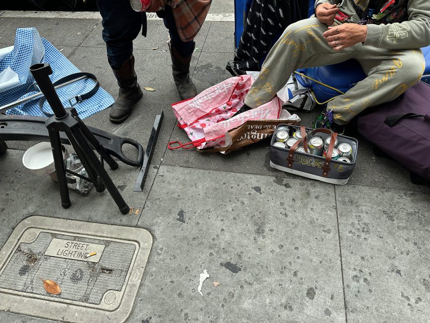 Two people sit on a city sidewalk surrounded by bags, a folding chair frame, and an open container of empty soda cans near a utility cover labeled "Street Lighting.
