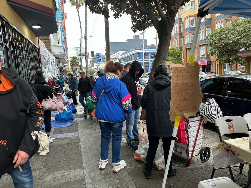 People gather on a city sidewalk near tables with bags and boxes; a handwritten menu is attached to a cart in the foreground.