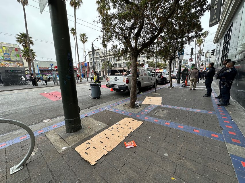 A city sidewalk with cardboard pieces on the ground, a tree, a parked pickup truck, police officers, and pedestrians in an urban setting.
