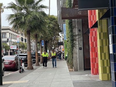 Two people in safety vests walk on a city sidewalk lined with palm trees, parked cars, and colorful tiled buildings. A scooter is parked near the curb.