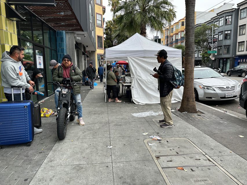 People stand near a white tent on a city sidewalk with parked cars, a motorcycle, and suitcases visible. Buildings and palm trees line the street.