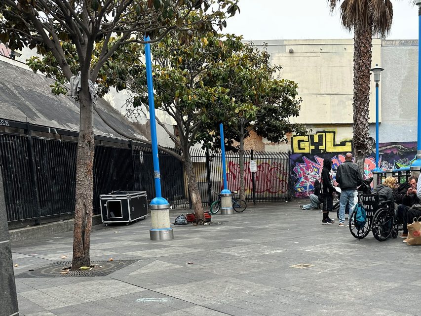 Urban sidewalk scene with trees, blue lampposts, graffiti on walls, a wheelchair, equipment case, and several people gathered near a palm tree.