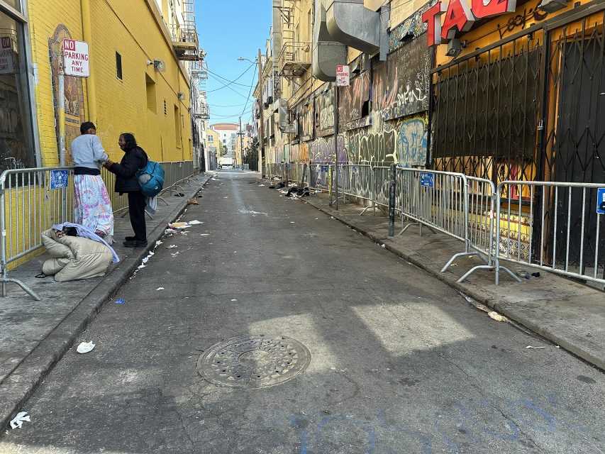 A narrow alley with metal barricades, graffiti-covered walls, scattered trash, and three people, two standing and one sitting on the ground near a yellow building.