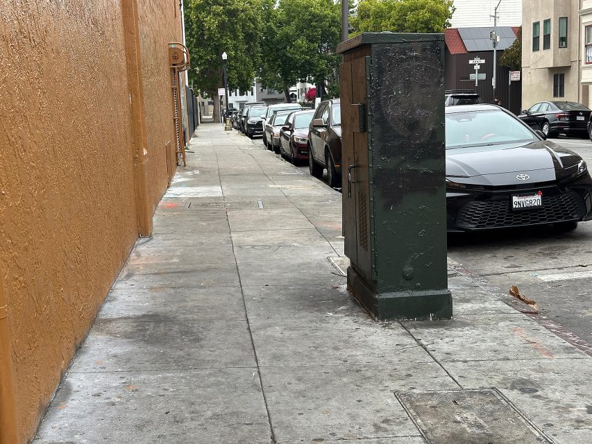 A city sidewalk with parked cars, an orange building on the left, and a large utility box near the curb. Trees and buildings line the street in the background.