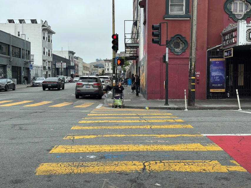 A person with a shopping cart waits at a crosswalk on a city street with buildings, cars, and traffic lights visible.