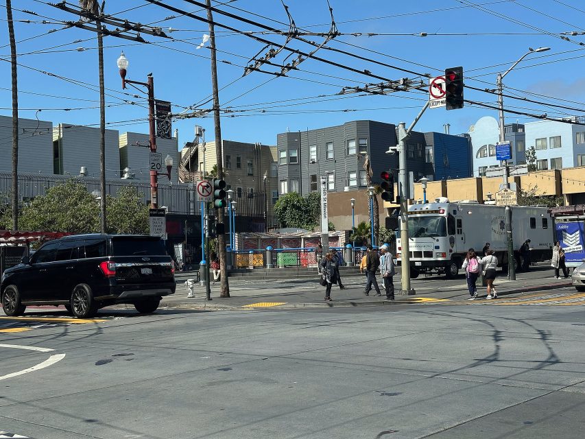 Urban street intersection with pedestrians crossing, vehicles waiting at a red light, overhead wires, and mid-rise buildings in the background under a clear blue sky.