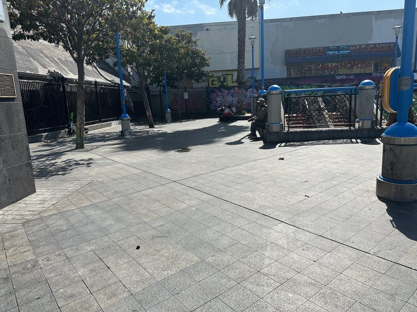 A paved urban plaza with blue lamp posts, trees, benches, and a mural-covered wall in the background on a sunny day.