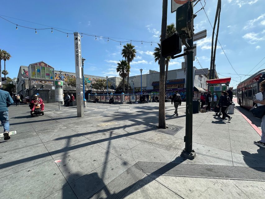 A busy urban street scene with people walking, palm trees, a colorful building, a scooter rider, and a red transit shelter under a clear sky.