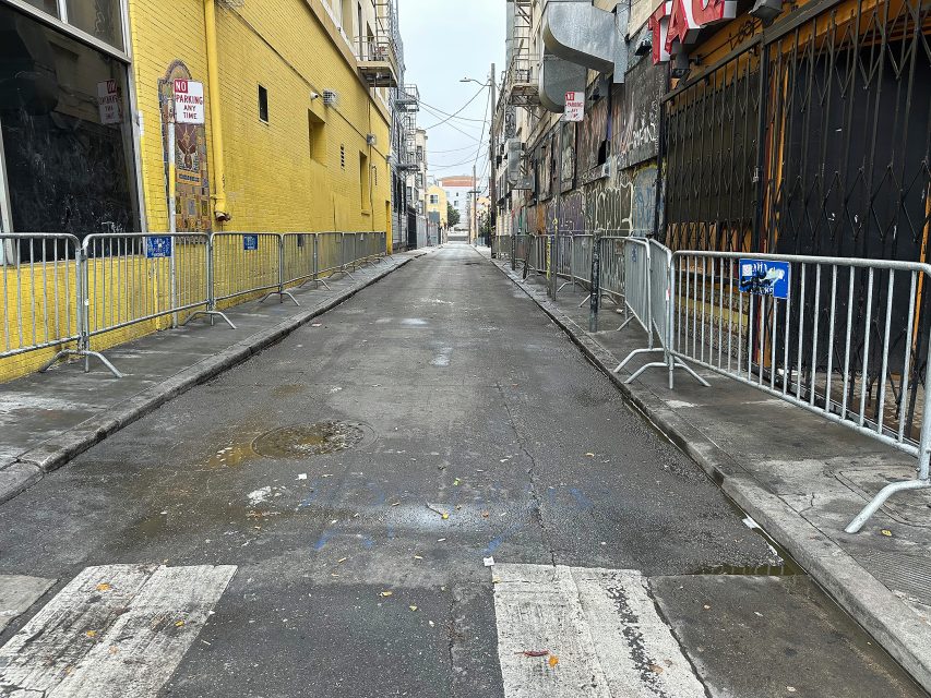 A deserted urban alleyway with metal barricades lining both sides, yellow and gray buildings, and a wet pavement under overcast skies.