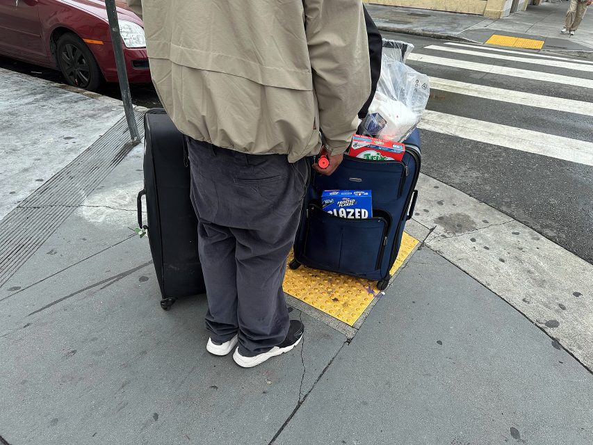 A person stands on a city sidewalk with two large suitcases, one blue and one black, near a crosswalk.