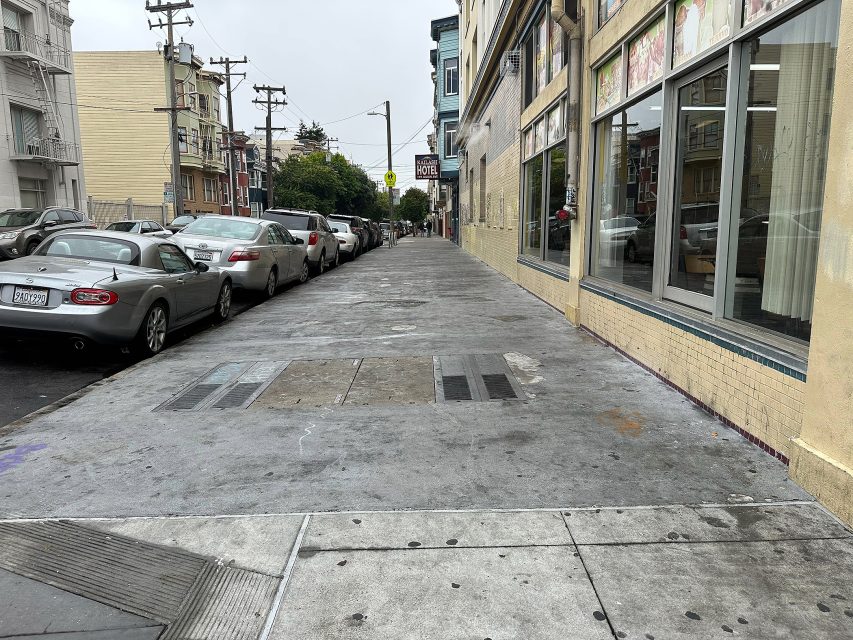 A city sidewalk runs alongside parked cars and a row of buildings on an overcast day. The street is empty of pedestrians.