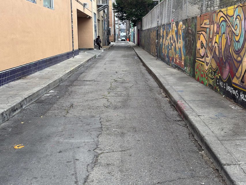 A narrow urban alley with cracked pavement, graffiti-covered walls on the right, and a tan building on the left. A few people are visible in the distance.