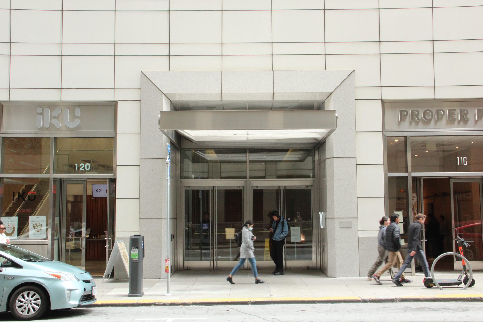 People walk past a modern building entrance with glass doors, flanked by storefronts named "IKU" and "Proper" on a city street.