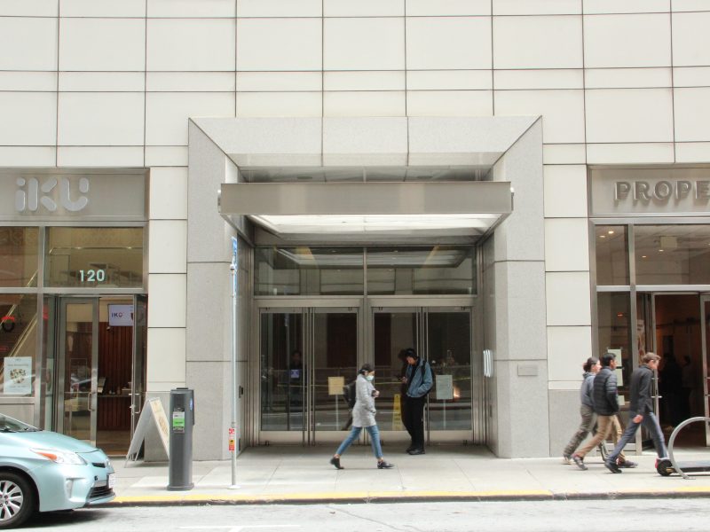 People walk past a modern building entrance with glass doors, flanked by storefronts named "IKU" and "Proper" on a city street.