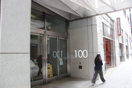 A person walks past the entrance of a building marked "100" on a city sidewalk, with a "Proper Food" storefront visible next door.