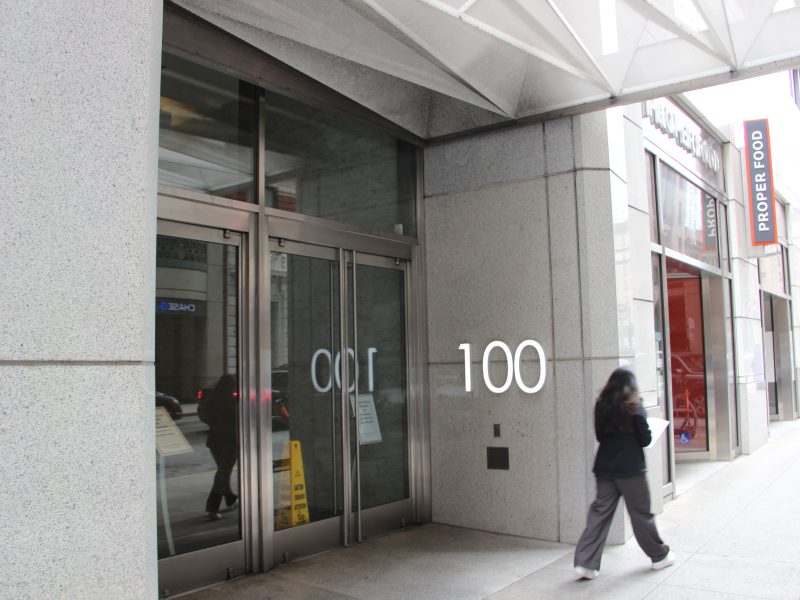 A person walks past the entrance of a building marked "100" on a city sidewalk, with a "Proper Food" storefront visible next door.