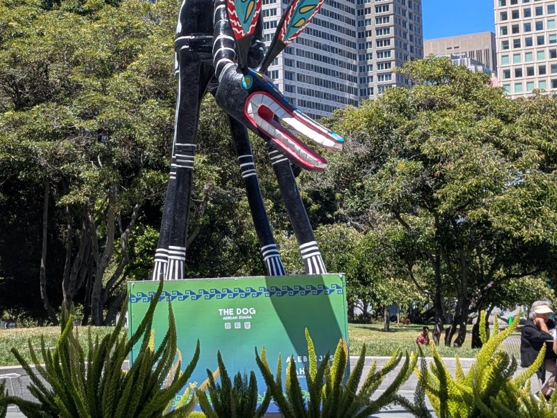 Large, colorful dog sculpture with long legs and patterned wings stands in an urban park, surrounded by trees, skyscrapers, and potted plants in the foreground.