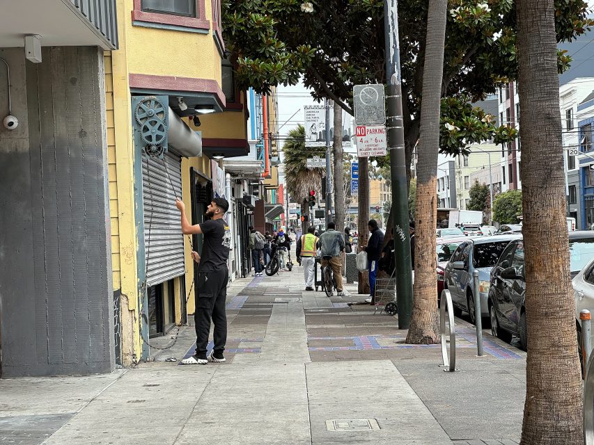 A person stands on a city sidewalk, reaching toward a security gate on a storefront; people, trees, and parked cars line the street.
