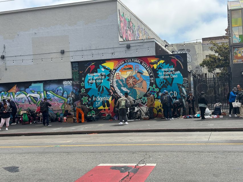 People gather on a city sidewalk in front of a mural reading "American Indian Cultural District" with colorful graffiti and text on the wall behind them.