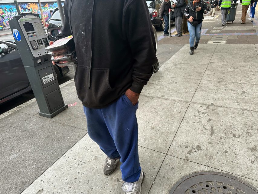 A person in a black hoodie and blue pants stands on a city sidewalk, holding food containers, with other people and cars visible in the background.