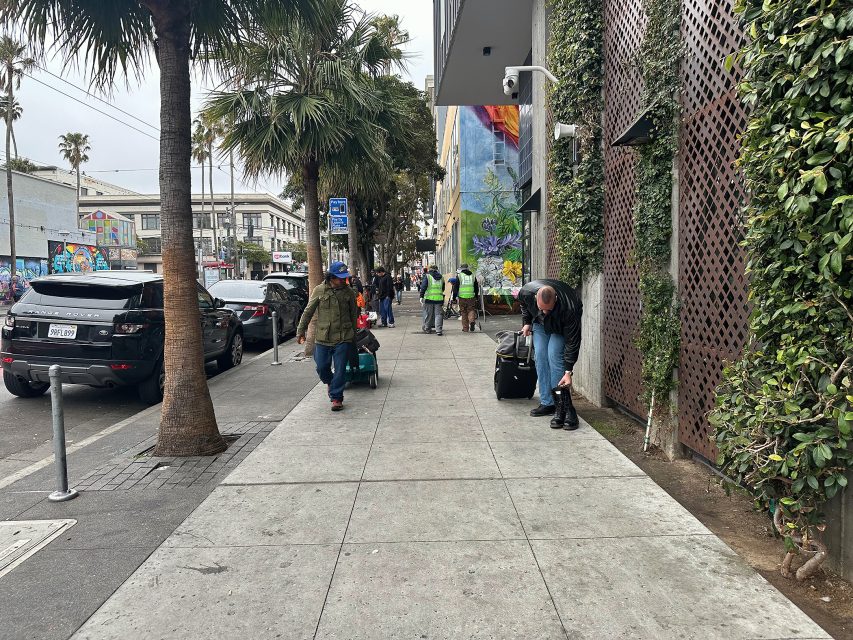 People walk along a city sidewalk lined with palm trees, parked cars, and street art; one person bends down near a suitcase on the right.