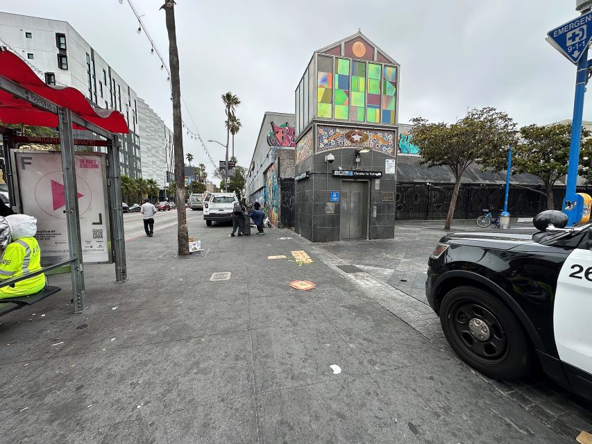 A city street corner with a colorful mural, a police car, two people talking, a person in a yellow vest, bus stop shelter, and various signs on the sidewalk.