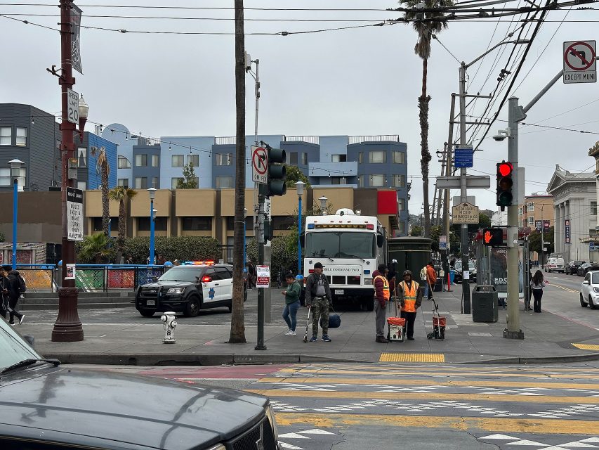 A city street scene shows police and public works employees near a truck on a corner, with pedestrians walking and a police car parked nearby.