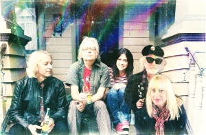 Five adults sit on steps outside a building, some holding drinks. One person wears sunglasses and a hat; another has a striped shirt. The image has a faded, vintage appearance.