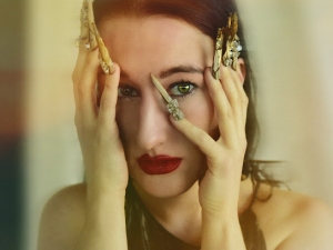 A person with red lipstick holds their face with both hands, wearing long, decorative fingernail extensions on their fingers.