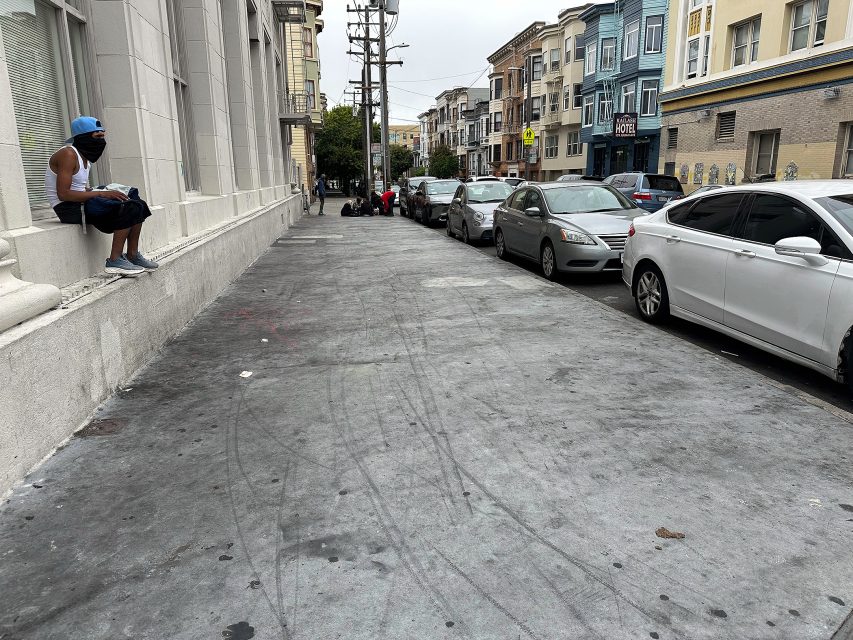 A man in a blue cap and mask sits on a ledge beside a wide sidewalk; cars are parked along the street with several people gathered in the distance.