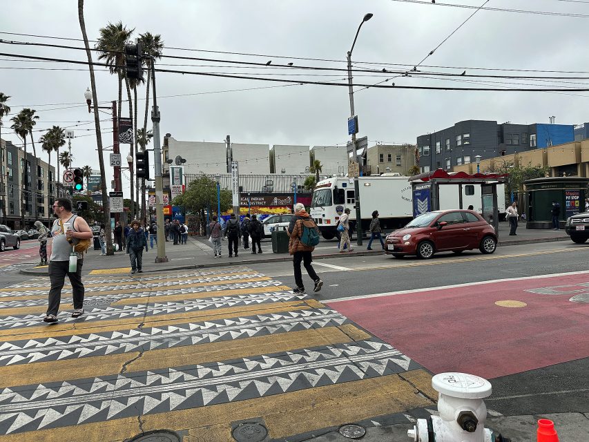 People cross a patterned crosswalk at an urban intersection with cars, palm trees, and buildings under a cloudy sky.