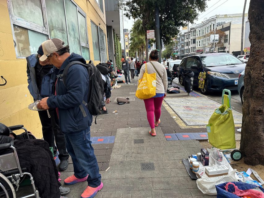 People stand and walk along a busy urban sidewalk lined with personal belongings, goods for sale, and shopping carts near parked cars and buildings.