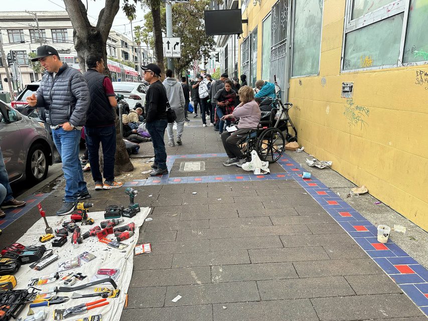 A group of people stand and sit along a busy city sidewalk; tools are laid out on a blanket, and some people appear to be waiting or resting.