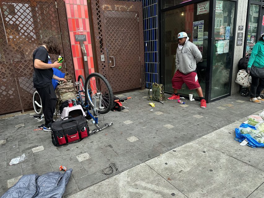 Two men stand on a littered sidewalk; one is working on a bicycle near bags and tools, while the other stands nearby in front of a storefront.
