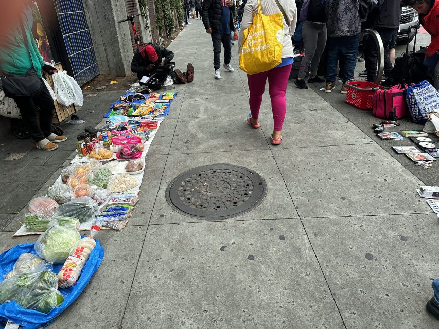 Vegetables, packaged foods, and other goods are displayed for sale on a city sidewalk as people walk and gather nearby.