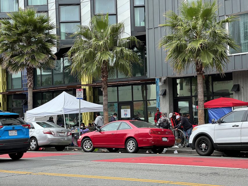 Several tents and people gather on a city sidewalk beside parked cars and palm trees in front of a modern building.