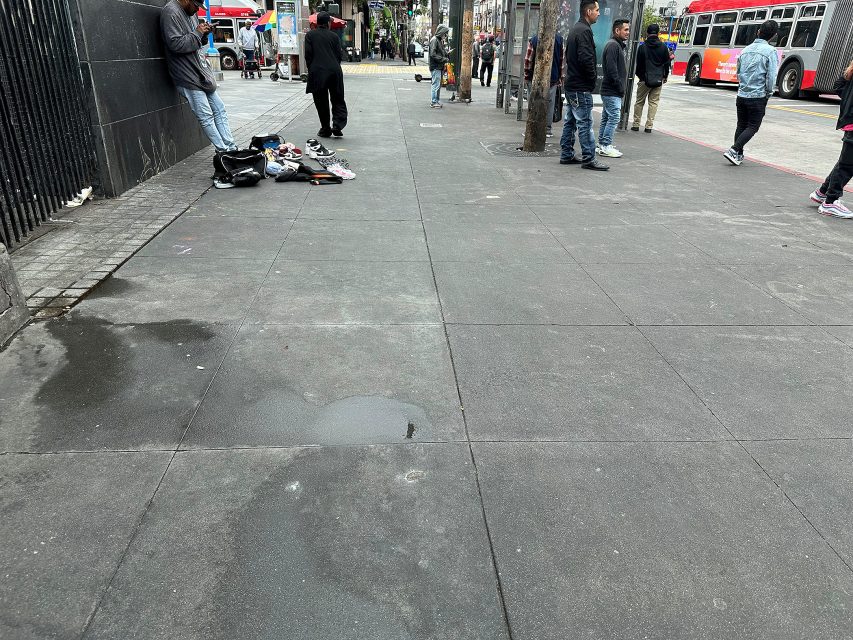 Several people stand and walk along a city sidewalk near a bus stop; items are laid out on the ground, and buses are visible in the background.