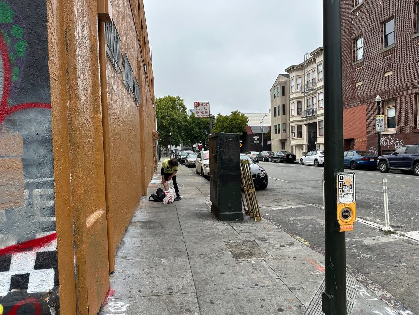 A person bends over near bags on a city sidewalk beside an orange building, with parked cars and street signs visible in the background.