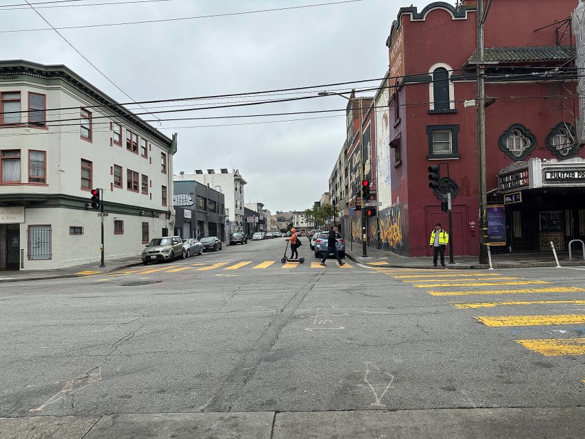 A city intersection with pedestrians crossing the street, cars stopped at a red light, and a security guard standing near a building with graffiti.