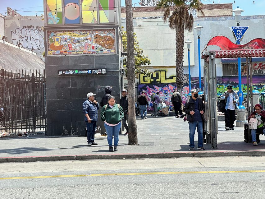 People wait at a city bus stop surrounded by graffiti-covered walls, with a palm tree and buildings in the background on a sunny day.