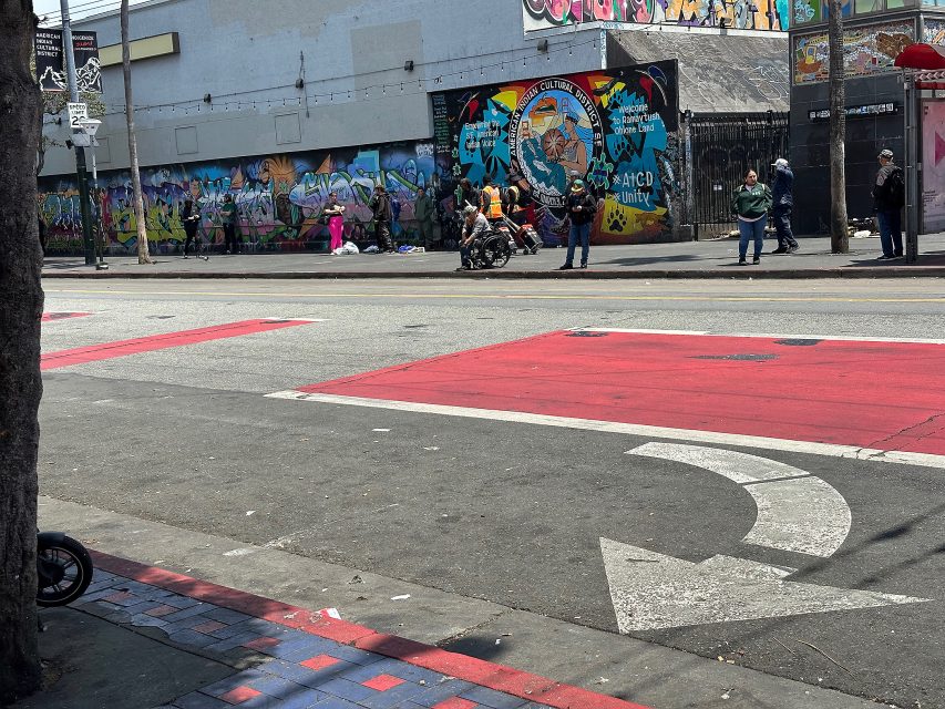 People stand and sit along a city street with colorful graffiti murals on a building in the background. A person in a wheelchair is in the center of the scene.